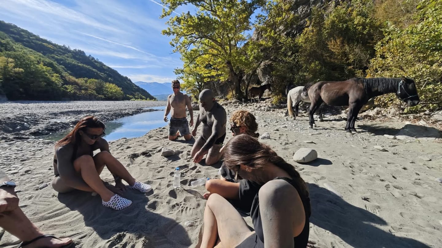 Riders resting on a riverbank while the horses wait